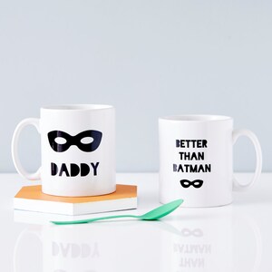 Peut inclure: Deux tasses en c&eacute;ramique blanche avec du texte noir et un motif de masque noir. La tasse de gauche dit "Daddy" et la tasse de droite dit "Better Than Batman".