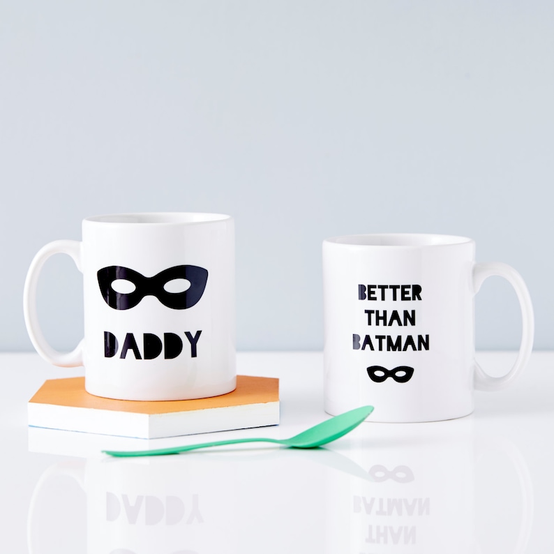 Peut inclure: Deux tasses en c&eacute;ramique blanche avec du texte noir et un motif de masque noir. La tasse de gauche dit "Daddy" et la tasse de droite dit "Better Than Batman".