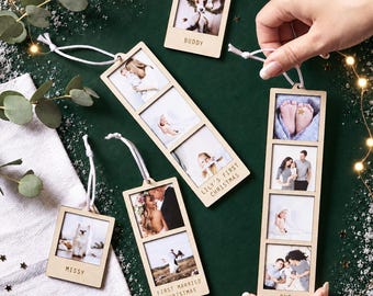 May include: A collection of light-colored wooden photo ornaments with white hanging strings. The ornaments feature square photo inserts and engraved text including "BUDDY," "LILY'S FIRST CHRISTMAS," "FIRST MARRIED CHRISTMAS," and "OUR FAMILY."