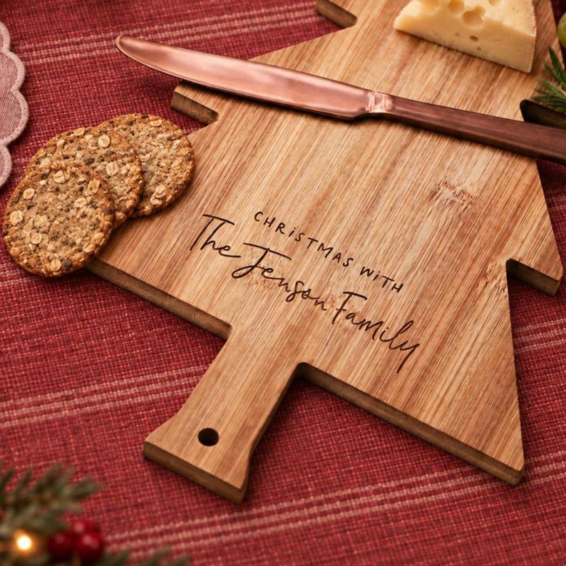 Pode incluir: Uma t&aacute;bua de servir de bambu em forma de &aacute;rvore gravada com o texto "CHRISTMAS WITH The Jenson Family", com uma faca de queijo cor de cobre por cima, acompanhada por tr&ecirc;s bolachas redondas texturizadas e um peda&ccedil;o de queijo branco.