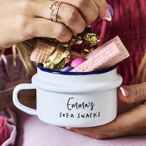 Pode incluir: Caneca de esmalte branca com borda azul, cheia de lanches. A caneca tem uma alça e as palavras "Emma's SOFA SNACKS" em escrita preta. A caneca é preenchida com wafers rosa, doces embalados e doces coloridos.