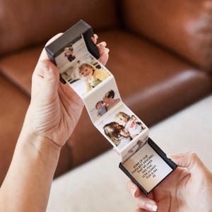 May include: A black square gift box containing an accordion-style pull-out photo strip. The strip features multiple colour photographs of children and a printed message that reads: SOME OF OUR FAVOURITE MEMORIES THAT WE MADE THIS YEAR, LOVE MUMMY, ELSA & MIA XX.
