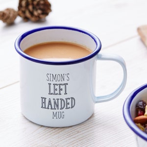 Może przedstawiać: Biały emaliowany kubek z niebieską obw&oacute;dką i uchwytem, wypełniony jasno brązową cieczą. Kubek ma czarny napis "SIMON'S LEFT HANDED MUG". Szyszka sosnowa w tle.