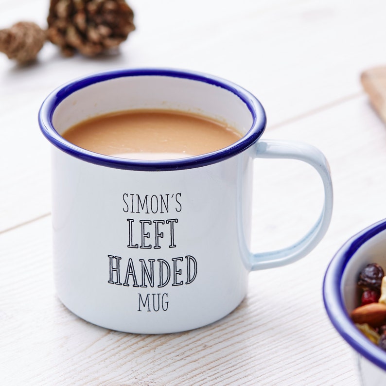 Może przedstawiać: Biały emaliowany kubek z niebieską obw&oacute;dką i uchwytem, wypełniony jasno brązową cieczą. Kubek ma czarny napis "SIMON'S LEFT HANDED MUG". Szyszka sosnowa w tle.