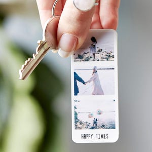 Hand holding a Personalised Photo Strip Keyring featuring three custom photo slots and the caption 'HAPPY TIMES'; a Custom Photo Gift Keyring and Personalized Photo Memory Keychain attached to a silver key against a soft background with greenery.