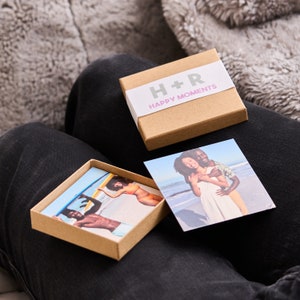 Puede incluir: Una caja marr&oacute;n con el texto "H+R Happy Moments" en la parte superior. La caja contiene dos peque&ntilde;as impresiones fotogr&aacute;ficas. Una foto muestra una pareja abraz&aacute;ndose en una playa, y la otra muestra a una persona surfeando.