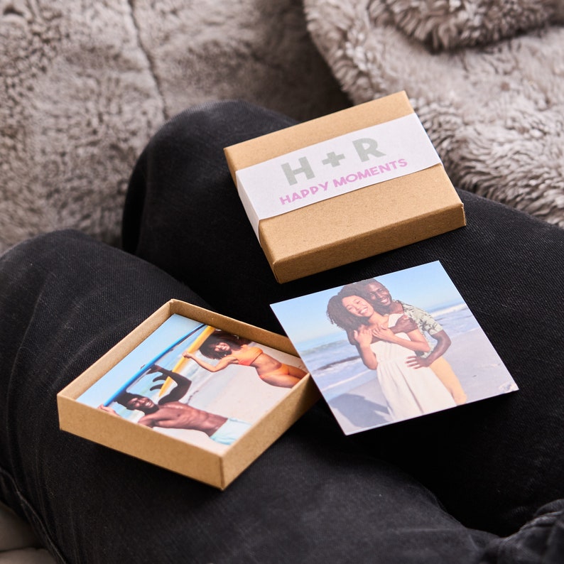 Puede incluir: Una caja marr&oacute;n con el texto "H+R Happy Moments" en la parte superior. La caja contiene dos peque&ntilde;as impresiones fotogr&aacute;ficas. Una foto muestra una pareja abraz&aacute;ndose en una playa, y la otra muestra a una persona surfeando.