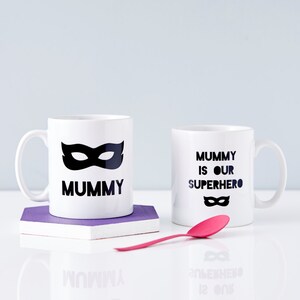 Peut inclure: Deux tasses en c&eacute;ramique blanche avec du texte noir et un motif de masque noir. La tasse de gauche dit "Mummy" et la tasse de droite dit "Mummy is our superhero".