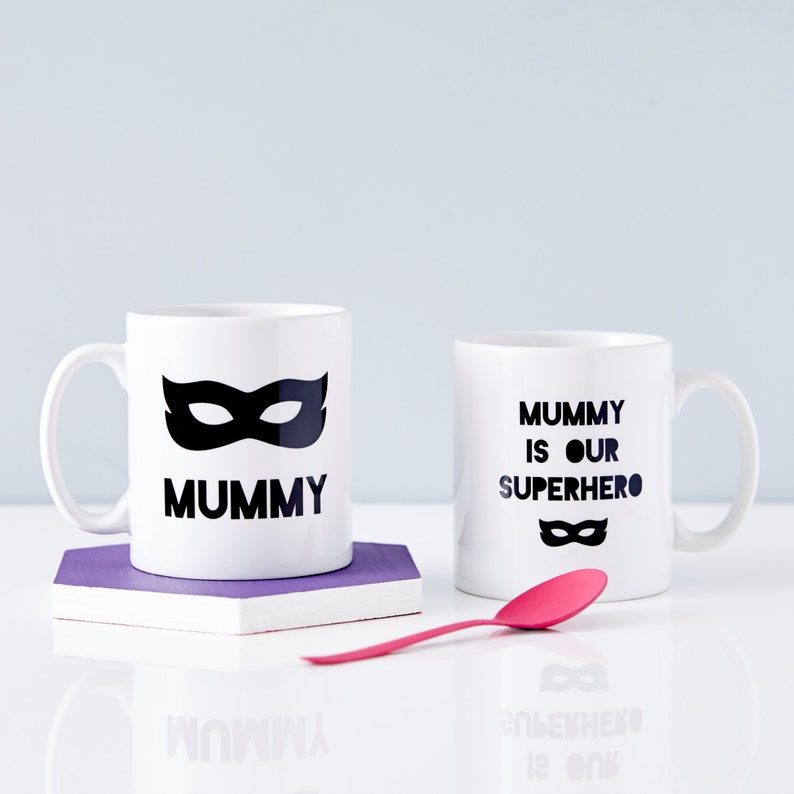 Peut inclure: Deux tasses en c&eacute;ramique blanche avec du texte noir et un motif de masque noir. La tasse de gauche dit "Mummy" et la tasse de droite dit "Mummy is our superhero".