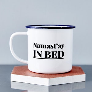 Op de afbeelding: Witte emaille mok met een blauwe rand en oor. De mok heeft de tekst "Namast'ay IN BED" in het zwart, rustend op een geometrisch, roodbruin en wit oppervlak. Een leuk cadeau voor thee- of koffiedrinkers.