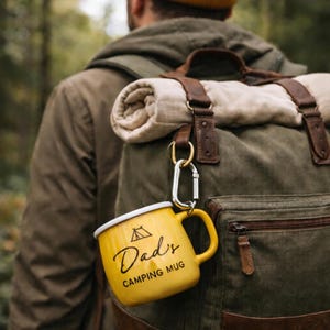 May include: Yellow camping mug with the text "Dad's Camping Mug" and a small tent graphic. The mug is attached to a green backpack with a rolled-up blanket. The mug has a white rim and a yellow handle.