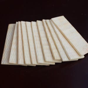 Könnte beinhalten: Eine Ansammlung hellfarbiger Holzbretter, fächerförmig angeordnet. Die Bretter haben eine natürliche Holzmaserung und sind rechteckig. Der Hintergrund ist eine dunkle, einfarbige Farbe.