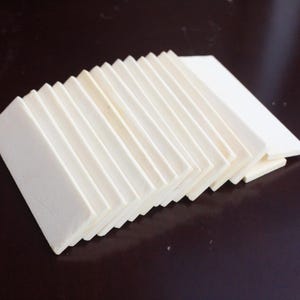 Puede incluir: Una pila de hojas de papel rectangulares de color blanquecino, dispuestas en forma de acordeón. Las hojas están dispuestas en forma de abanico, revelando sus bordes. El papel tiene una superficie texturizada y está sobre un fondo marrón oscuro.