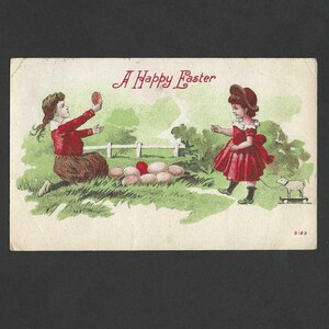 May include: Vintage Easter postcard featuring two young girls in red dresses and a lamb on wheels. The text "A Happy Easter" is written in red script above the girls. The background shows a green field, a white fence, and a collection of eggs.