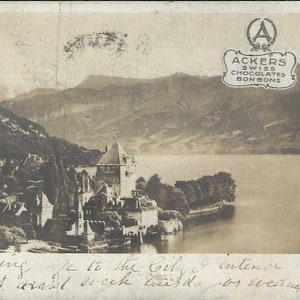 Peut inclure: Une carte postale vintage en noir et blanc représentant un château sur un lac avec des montagnes en arrière-plan. La carte postale porte un timbre et un logo pour Ackers Swiss Chocolates Bonbons.