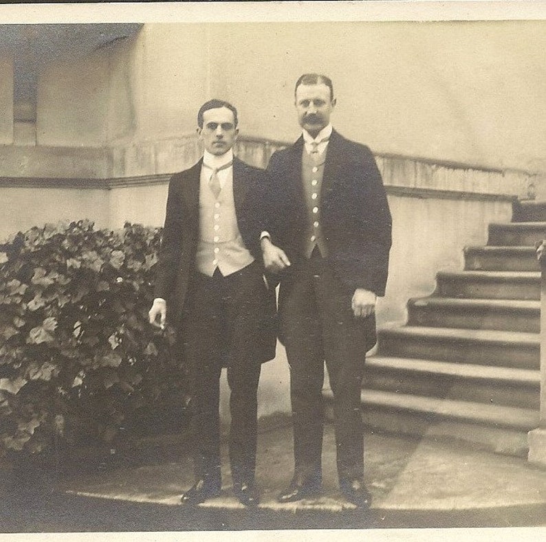 May include: Two men in formal attire stand in front of a staircase. They are both wearing suits, vests, and ties. The man on the left is holding a hat in his hand. The man on the right has a mustache.