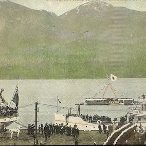 May include: A black and white photograph showing two ships docked at a pier. The vessels are decorated with flags and bunting. People can be seen on the decks of the ships and on the pier. The ships are in front of a mountain range.
