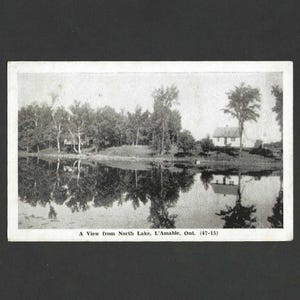 May include: Black and white vintage postcard featuring a scenic view of a lake with trees and a small house on the shore. The water reflects the trees and the house. Text at the bottom reads "A View from North Lake, L'Amable, Ont. (47-15)".