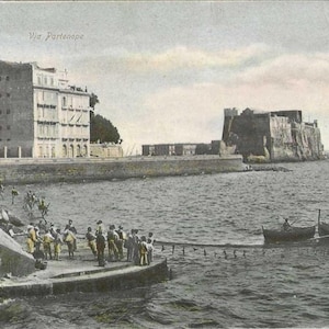 May include: A vintage postcard showing a view of the waterfront in Naples, Italy. The postcard features a building with the text "Via Partenope" at the top, a group of people standing on a pier, a boat in the water, and a large stone fortress in the distance.