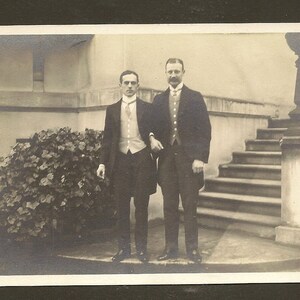 May include: Two men in formal attire stand on a stone platform in front of a staircase. The man on the left is wearing a dark suit with a white vest and a bow tie. The man on the right is wearing a dark suit with a white shirt and a tie. Both men are smiling.