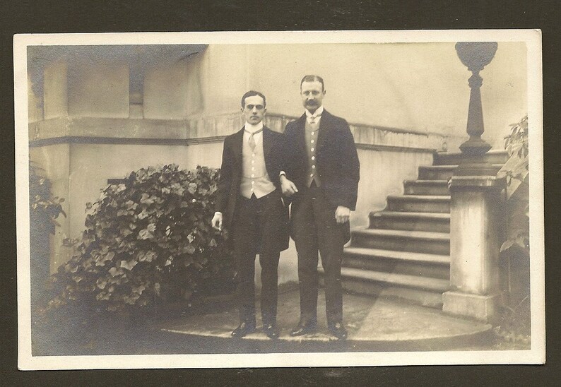 May include: Two men in formal attire stand on a stone platform in front of a staircase. The man on the left is wearing a dark suit with a white vest and a bow tie. The man on the right is wearing a dark suit with a white shirt and a tie. Both men are smiling.