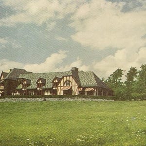 May include: A large, white and brown, multi-level house with a gabled roof and a green lawn in front. The house has a large front porch and several windows. There are trees in the background.