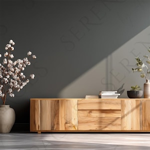 Op de afbeelding: Een houten kast met een natuurlijke afwerking, met een centrale lade en twee deuren. De kast is versierd met decoratieve items, waaronder boeken, een kleine vetplant en een vaas met groen. Een grote vaas met katoenstelen staat links.