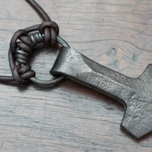 May include: A dark gray, hand-forged metal hammer pendant with a textured, rustic finish. The pendant is attached to a dark brown leather cord by a circular metal ring, secured with a series of intricate leather knots and wraps.