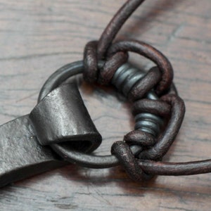 May include: A dark brown leather cord knotted around a circular dark gray metal ring. The metal ring features a textured, hammered surface and is attached to a flat, curved piece of dark gray metal with a matte finish.
