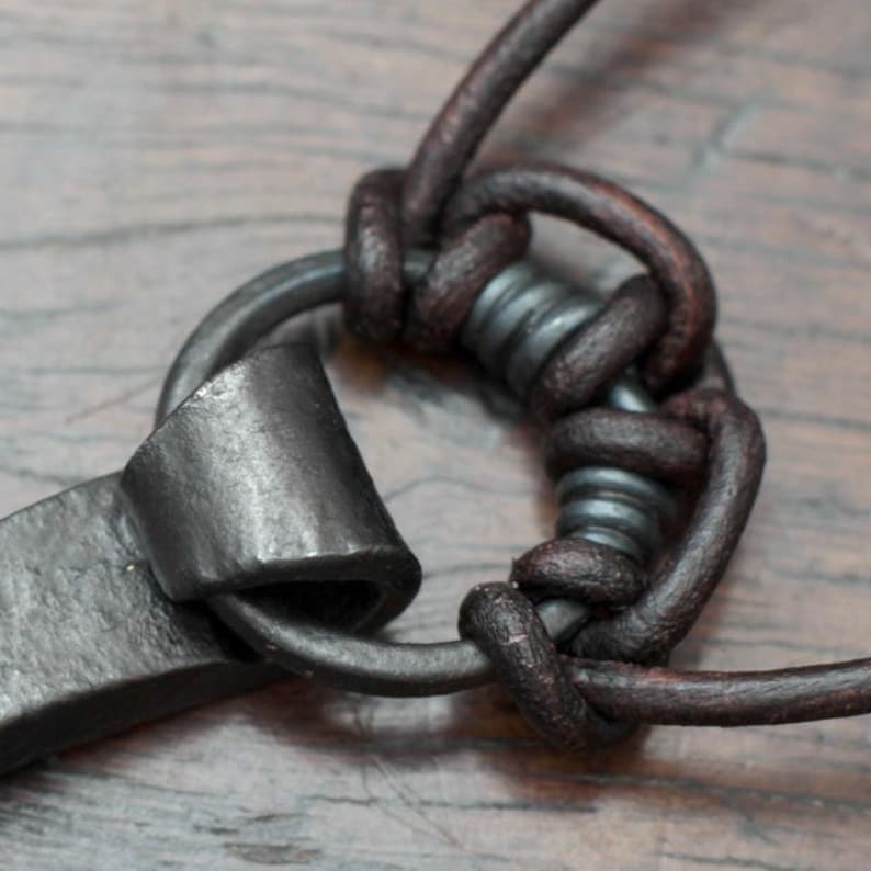 May include: A dark brown leather cord knotted around a circular dark gray metal ring. The metal ring features a textured, hammered surface and is attached to a flat, curved piece of dark gray metal with a matte finish.