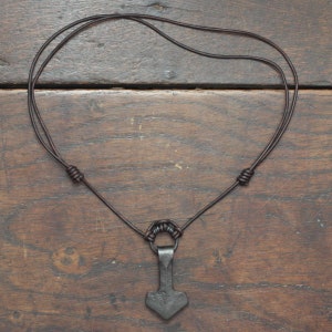 May include: A dark gray metal hammer-shaped pendant suspended from a thin dark brown leather cord. The cord features two adjustable sliding knots on either side and a small metal ring connecting the pendant to the leather necklace.