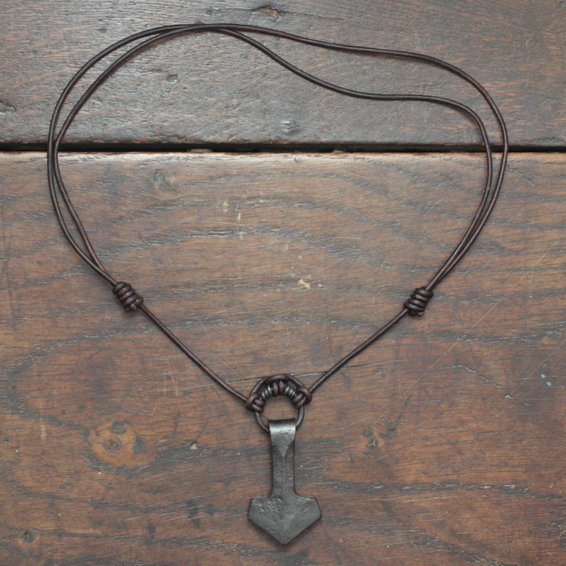 May include: A dark gray metal hammer-shaped pendant suspended from a thin dark brown leather cord. The cord features two adjustable sliding knots on either side and a small metal ring connecting the pendant to the leather necklace.