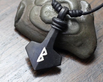 May include: A black metal pendant shaped like a hammer, featuring a silver-colored rune symbol on the front. The pendant is suspended from a black cord with decorative knots and rests against a smooth, grey, rounded stone surface.