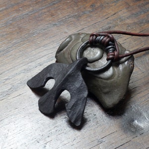Könnte beinhalten: Ein dunkelgrauer, handgeschmiedeter Metallanhänger in Form eines fliegenden Vogels. Der Anhänger hängt an einem dunkelbraunen Lederband an einem kreisförmigen Metallring, der mit einem gewickelten Lederknotendetail befestigt ist.
