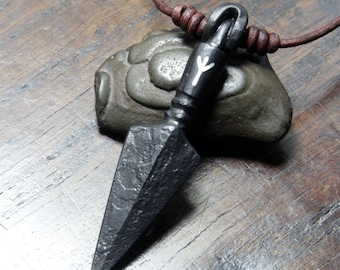 May include: A black forged metal pendant shaped like a spearhead, featuring a white Algiz rune symbol on the neck. The pendant hangs from a brown leather cord with small brown beads, showcasing a textured, dark, and rustic finish.