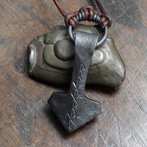 May include: A dark gray, hand-forged metal pendant shaped like a hammer, featuring etched runic symbols on its surface. The pendant hangs from a dark brown leather cord secured by a metal ring and a series of decorative knots.