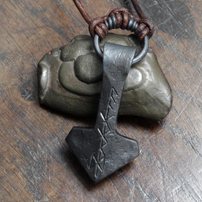 May include: A dark gray, hand-forged metal pendant shaped like a hammer, featuring etched runic symbols on its surface. The pendant hangs from a dark brown leather cord secured by a metal ring and a series of decorative knots.