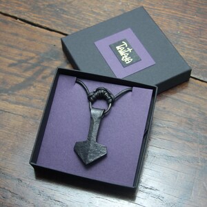 May include: A black metal hammer-shaped pendant on a black cord necklace, presented in a black gift box with a purple insert. The box lid features a purple square with the text "TAMAYA" in white lettering, resting on a wooden surface.