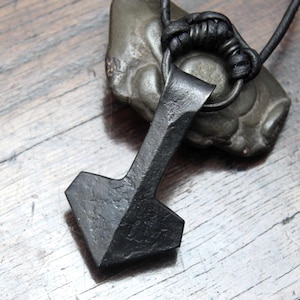 May include: A hand-forged black iron pendant shaped like a hammer, featuring a textured, rustic surface. The pendant is suspended from a black leather cord that passes through a circular metal ring at the top of the hammer-shaped piece.