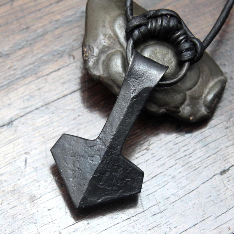 May include: A hand-forged black iron pendant shaped like a hammer, featuring a textured, rustic surface. The pendant is suspended from a black leather cord that passes through a circular metal ring at the top of the hammer-shaped piece.