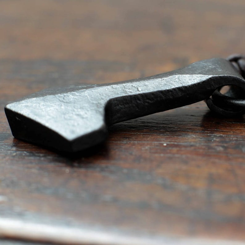 May include: A dark gray, hand-forged metal pendant shaped like a small axe head. The surface has a textured, hammered appearance with visible indentations and a matte finish. A small metal loop is attached to the top for a cord or chain.