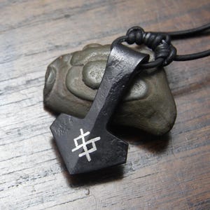 May include: A black metal pendant shaped like a hammer, featuring a white geometric symbol etched on the front. The pendant is attached to a black leather cord and rests against a smooth, grey, rounded stone with a textured surface.