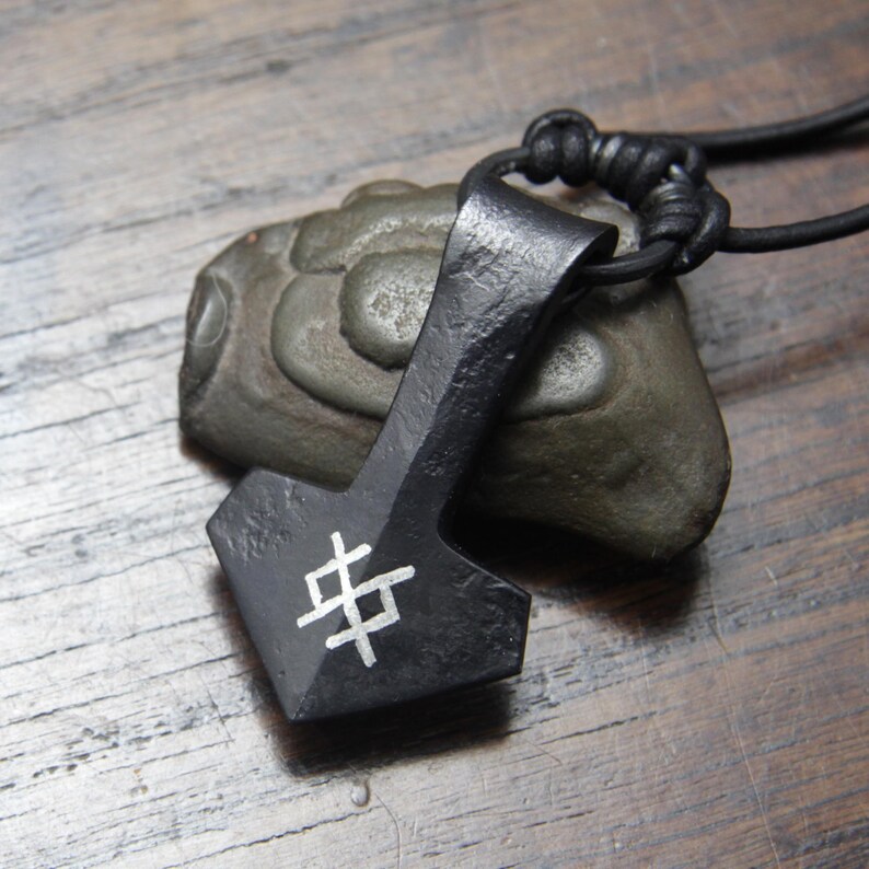 May include: A black metal pendant shaped like a hammer, featuring a white geometric symbol etched on the front. The pendant is attached to a black leather cord and rests against a smooth, grey, rounded stone with a textured surface.