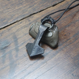 May include: A dark gray, hand-forged metal pendant shaped like a hammer, suspended from a thin black cord. The pendant features a textured, uneven surface and is positioned resting against a small, smooth, gray stone.