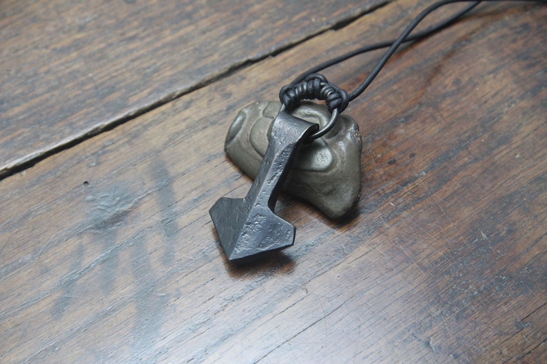 May include: A dark gray, hand-forged metal pendant shaped like a hammer, suspended from a thin black cord. The pendant features a textured, uneven surface and is positioned resting against a small, smooth, gray stone.