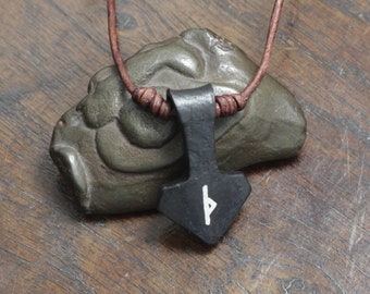 May include: A black forged metal pendant shaped like a hammer, featuring a white rune symbol on its front surface. The pendant is suspended from a thin brown leather cord with small brown leather knots on either side of the metal piece.