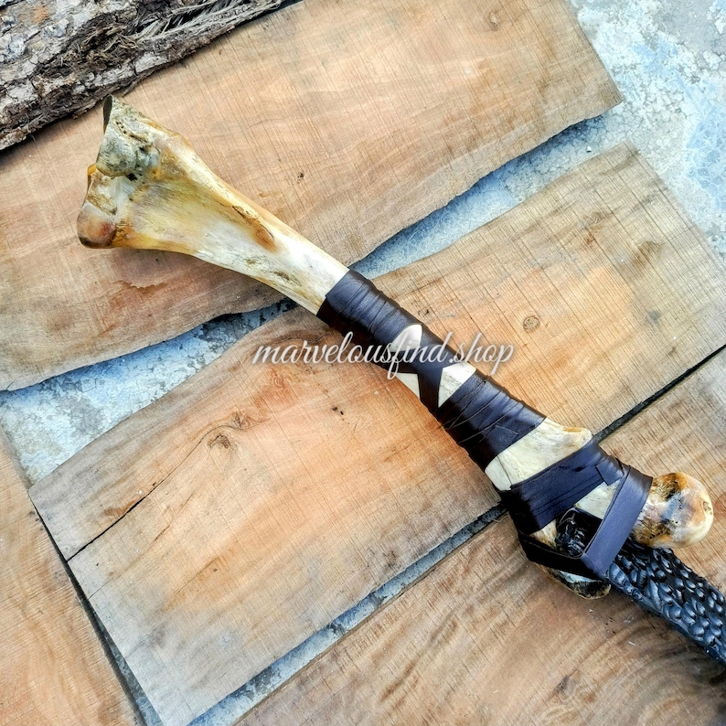May include: A handcrafted bone-handled weapon with a dark leather-wrapped grip and a braided handle. The bone has natural color variations, and the handle features a decorative pattern. The text "marvelousandshop" is visible.