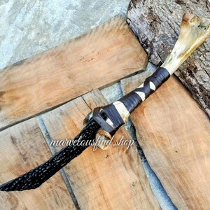 May include: A handcrafted dagger with a bone handle and a black, carved blade. The handle is wrapped with black and white leather. The text "marvelousfind shop" is visible on the wooden surface.