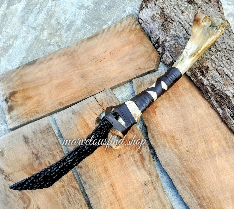 May include: A handcrafted dagger with a bone handle and a black, carved blade. The handle is wrapped with black and white leather. The text "marvelousfind shop" is visible on the wooden surface.