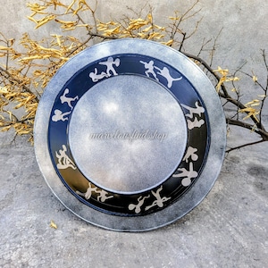 Op de afbeelding: Een zilverkleurige decoratieve plaat met een zwarte rand met witte silhouetfiguren. De plaat heeft een verweerde afwerking en de tekst "marvelousfindshop" is zichtbaar. De plaat is rond en lijkt van metaal te zijn.